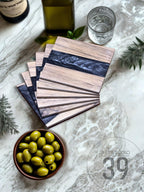 A stack of wooden coasters with a dark blue resin inlay. The coasters are made of light-colored wood and have a rectangular shape. A bowl of green olives and a glass of water are also visible.