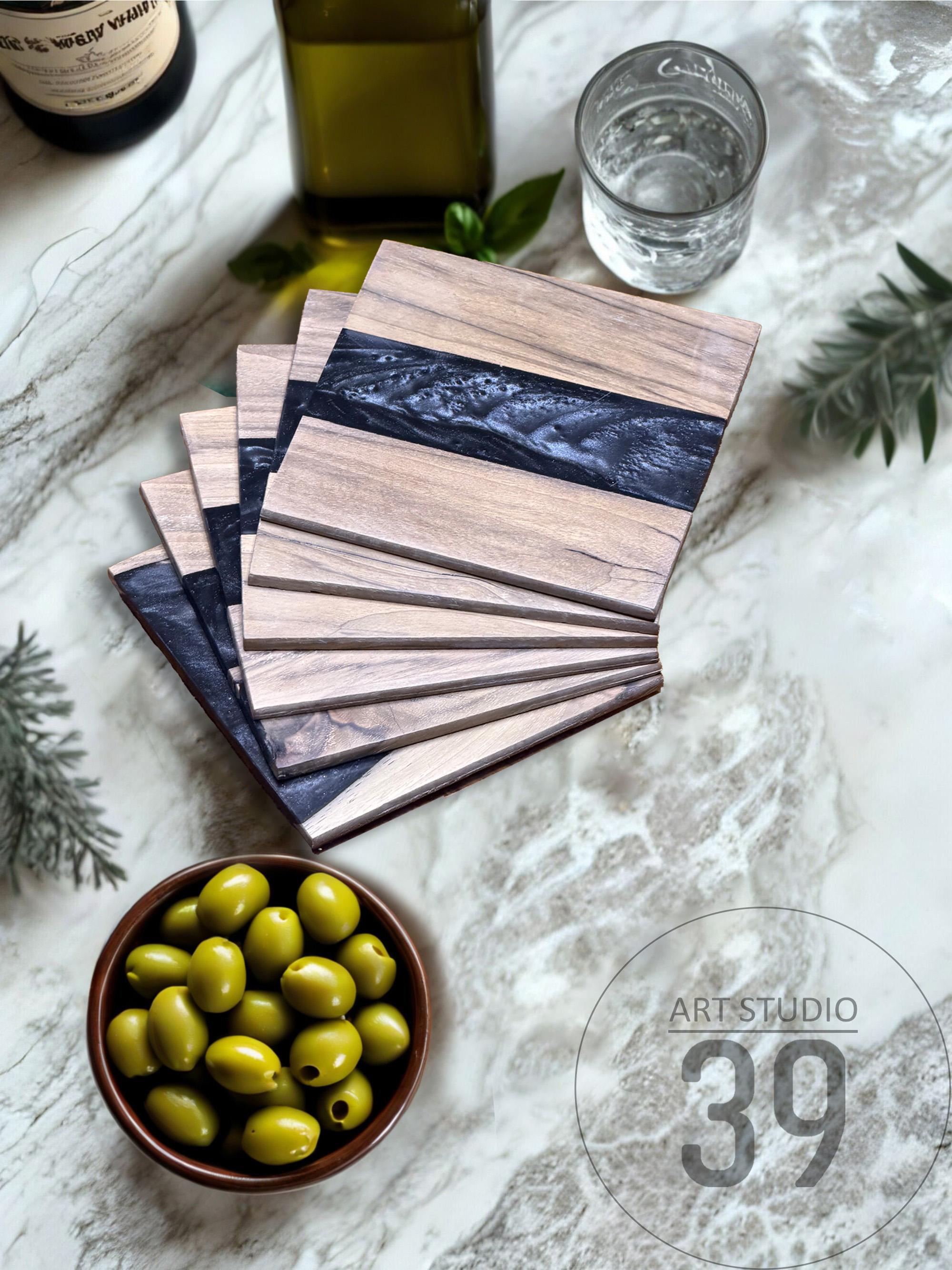 A stack of wooden coasters with a dark blue resin inlay. The coasters are made of light-colored wood and have a rectangular shape. A bowl of green olives and a glass of water are also visible.