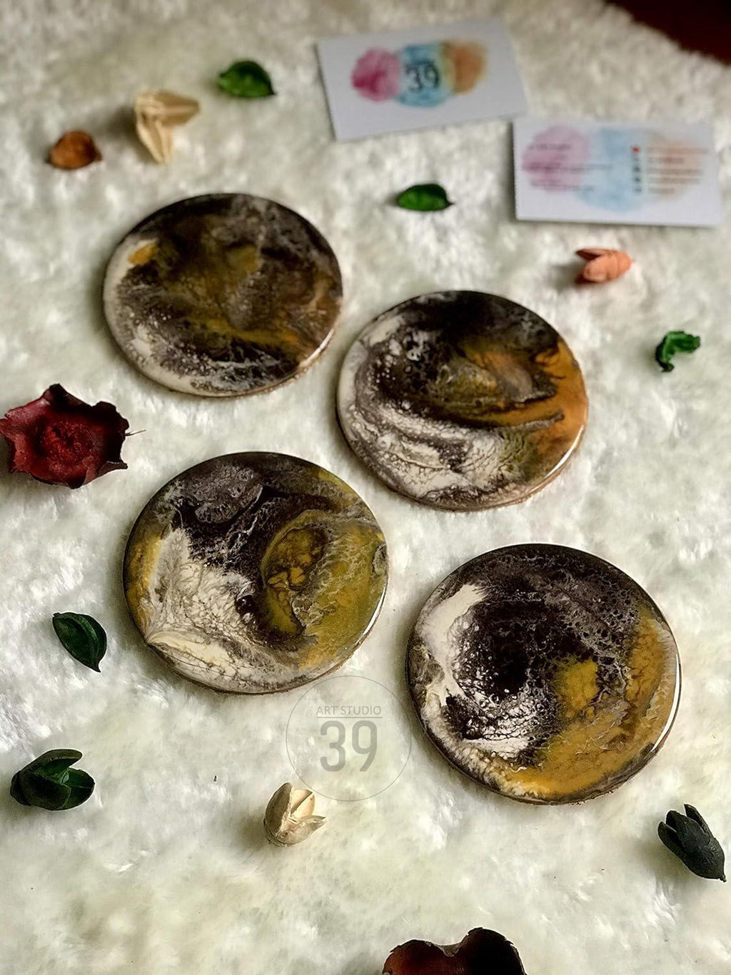 Four round coasters with abstract designs in shades of brown, gold, and white. The coasters are arranged on a white, textured surface, with decorative elements like leaves and small cards. The text "ART STUDIO 39" is visible.