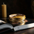 A stack of four round coasters with gold edges and a marbled design in shades of white, brown, and yellow. The coasters are resting on an open book. A textured, cylindrical candle is in the background.
