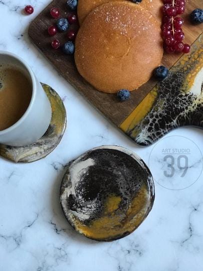 A white ceramic coffee cup with espresso sits on a decorative coaster with black, gold, and white swirls. A wooden cutting board holds pancakes and berries. A second coaster with similar design is also visible.