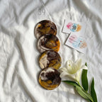 Four round coasters with a marbled design in shades of brown, gold, and white are stacked on a white surface. Two business cards with the text "Studio 39" and a white lily flower are also present.