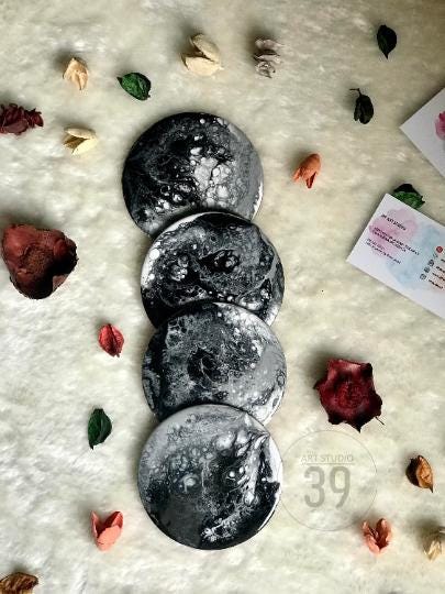 Four round coasters with a black and white marbled design are stacked on a white, textured surface. The coasters are surrounded by dried flowers and leaves. The bottom right coaster has the text "ART STUDIO 39".