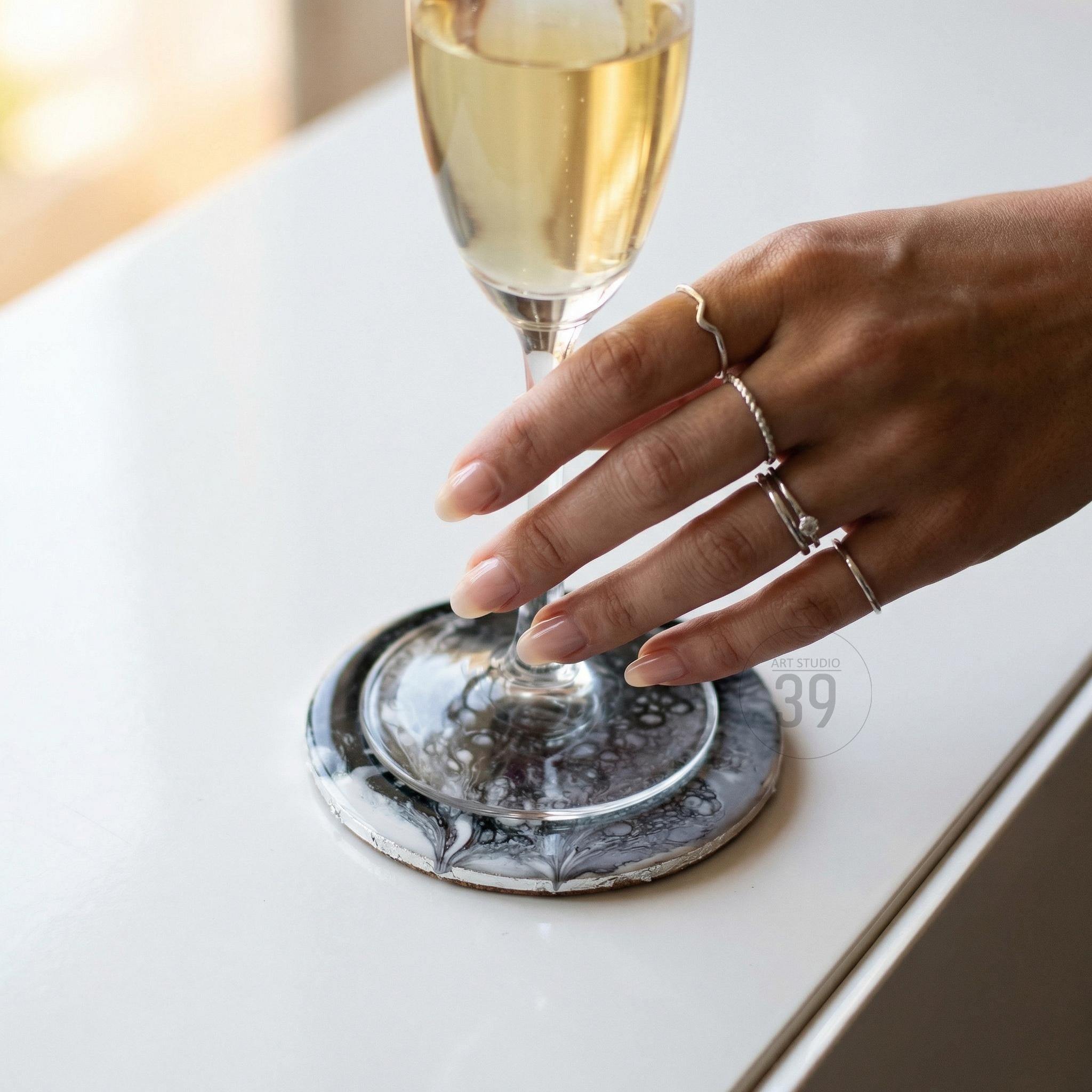 A hand wearing multiple silver rings holds a champagne glass above a round coaster. The coaster has a marbled black and white design. The glass is filled with a golden liquid.