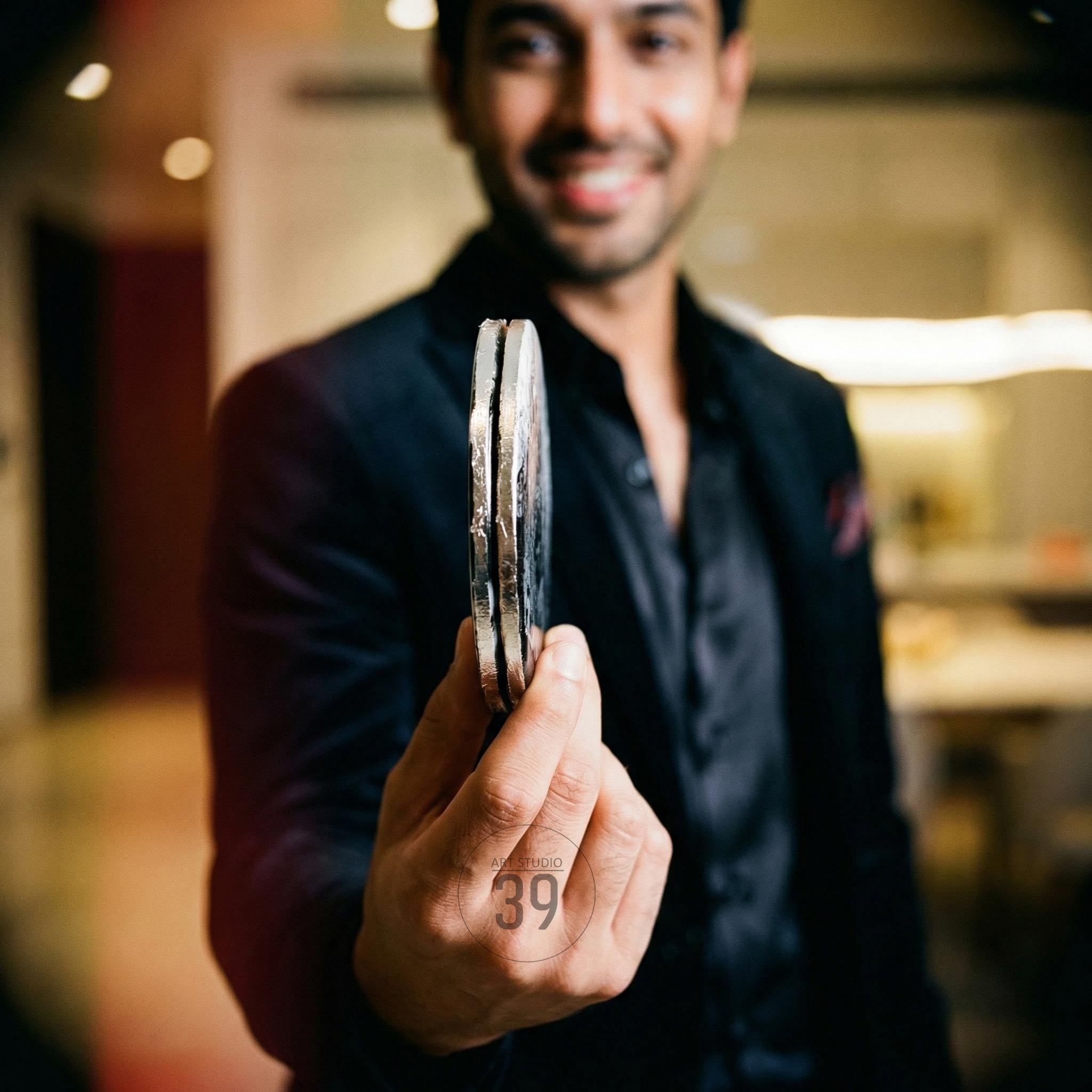 A person holding two silver-colored, circular objects. The objects have a textured edge and appear to be made of metal. The number "39" is printed on the person's hand. The person is wearing a black suit.
