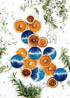 Overhead shot of a collection of dried orange slices and blue resin coasters with a white wave design. Green herbs and sprigs are scattered around the arrangement on a white surface. The image has a natural, organic aesthetic.