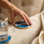 A round coaster with a blue and white ocean wave design and a gold rim. The coaster is being held by a hand wearing multiple gold rings and bracelets. The text "ART STUDIO 39" is visible.