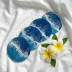 Four round coasters with a blue and white ocean wave design. The coasters are arranged on a white surface with two white and yellow plumeria flowers and green leaves. The coasters have the text "ART STUDIO 39".
