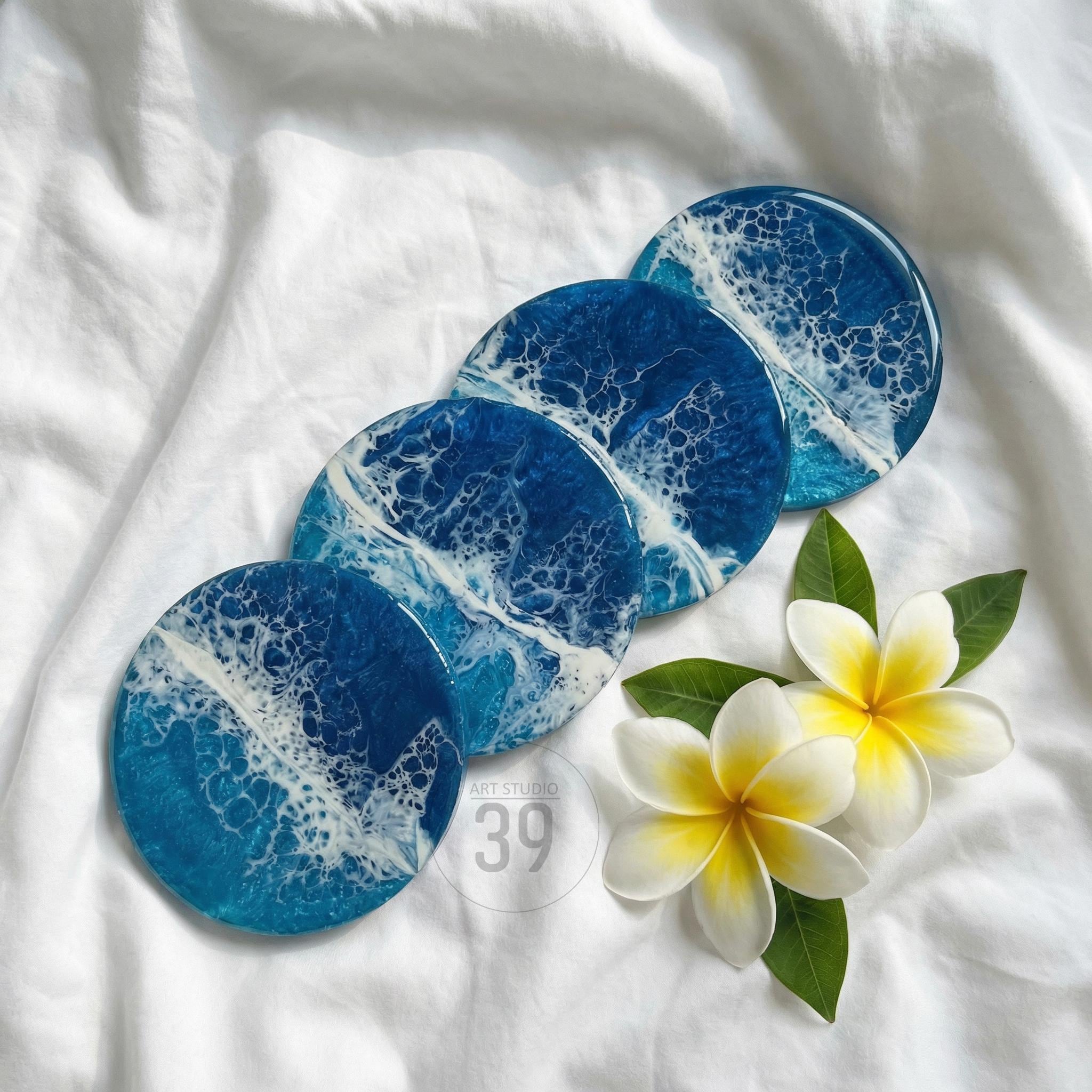 Four round coasters with a blue and white ocean wave design. The coasters are arranged on a white surface with two white and yellow plumeria flowers and green leaves. The coasters have the text "ART STUDIO 39".