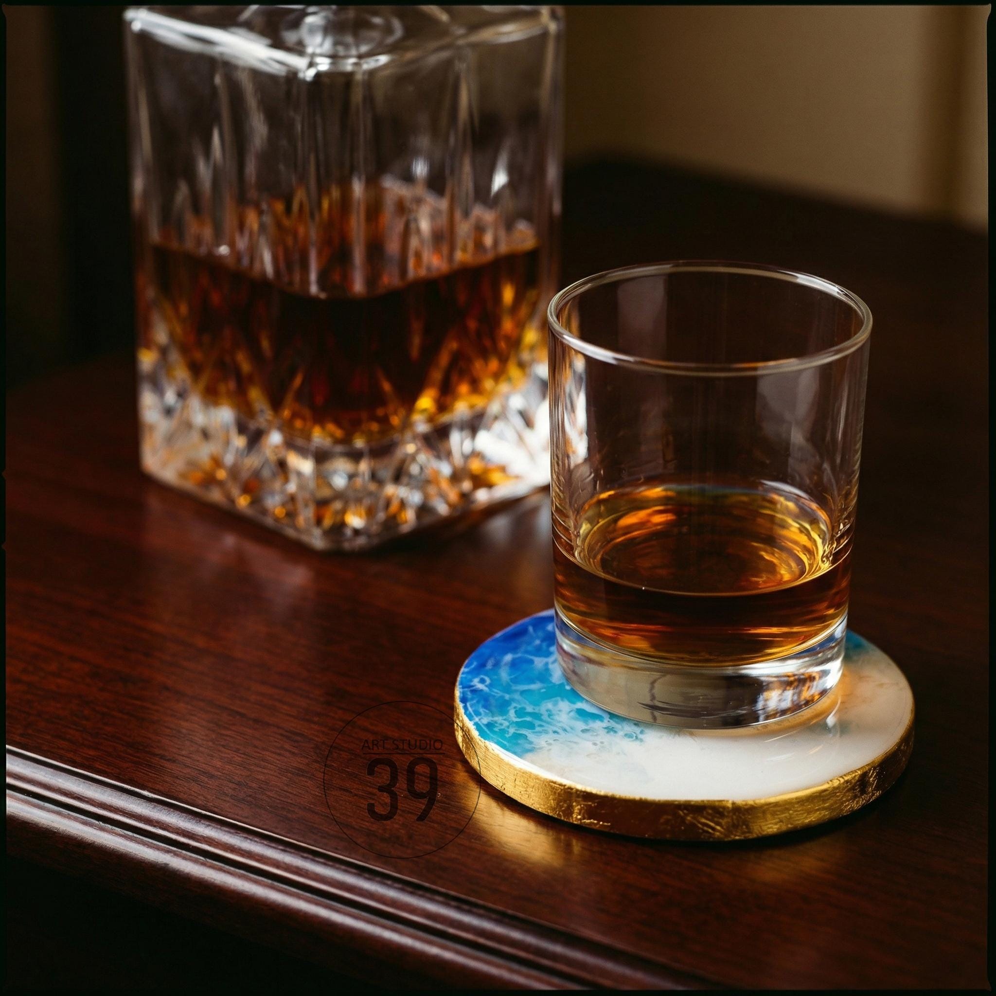 A rocks glass with amber liquid sits on a blue, white, and gold coaster. A crystal decanter filled with amber liquid is in the background. The coaster has a gold leaf edge and a blue and white ocean wave design.