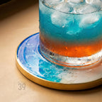 A refreshing cocktail in a clear glass with ice, resting on a round coaster. The drink has layers of blue and orange hues. The coaster features a blue and white marbled design with a gold edge. The number "39" is printed on the coaster.