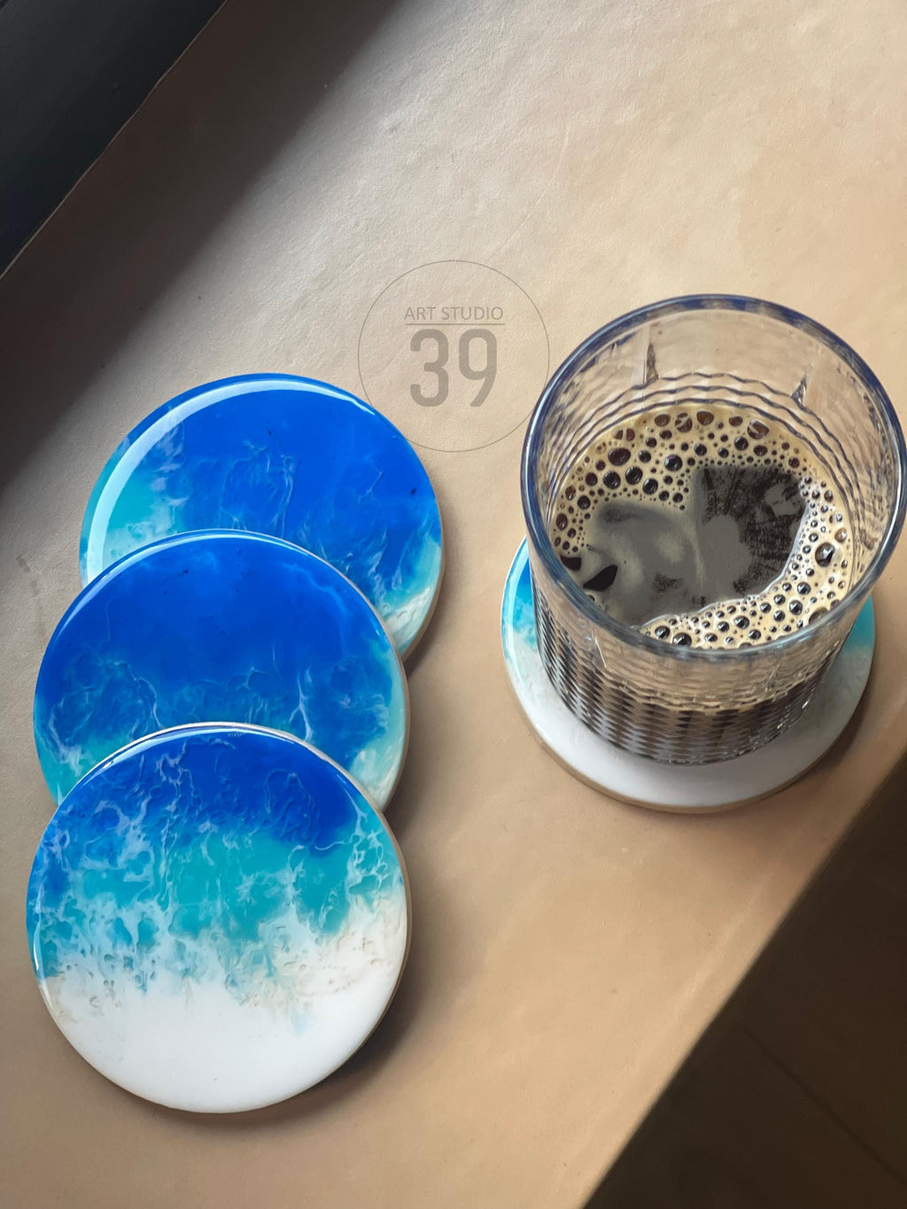 Set of four round coasters with a blue and white ocean wave design. A glass of coffee sits on one coaster. The coasters are made of resin and have a glossy finish. The image also shows the text "ART STUDIO 39".