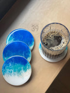 Set of four round coasters with a blue and white ocean wave design. A glass of coffee sits on one coaster. The coasters are made of resin and have a glossy finish. The image also shows the text "ART STUDIO 39".