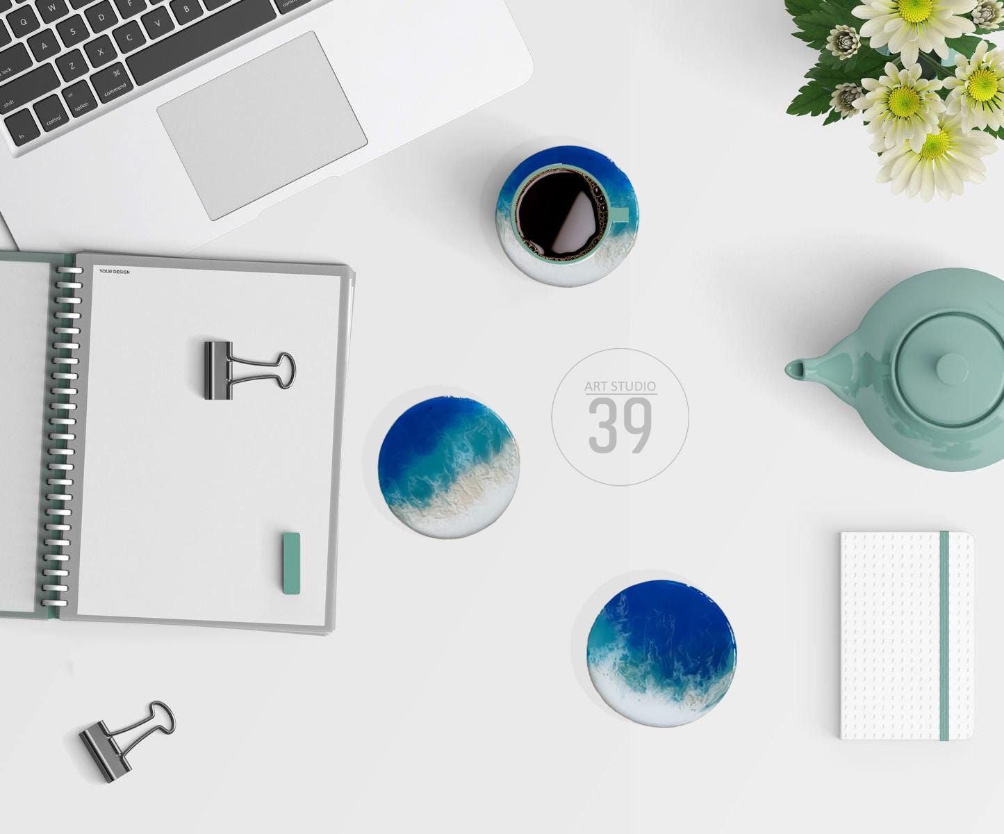 An overhead shot of a workspace with a laptop, notebook, and a cup of coffee. Two round coasters with blue and white designs are visible, along with a teapot and a small notepad. The text "ART STUDIO 39" is in the center.