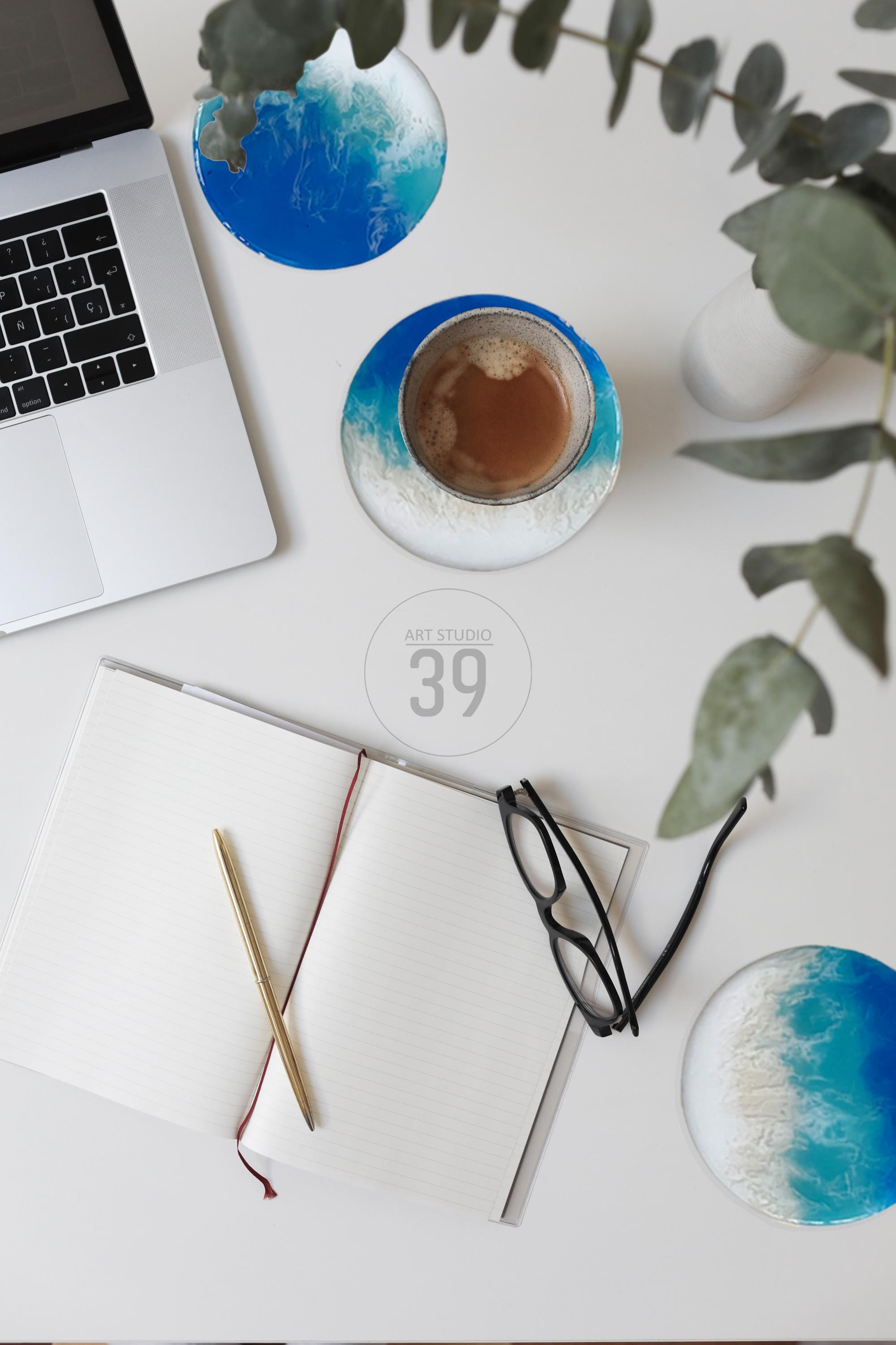 A top-down view of a workspace with a laptop, notebook, pen, and glasses. Two coasters with blue and white ocean wave designs are visible, one holding a cup of coffee. The text "ART STUDIO 39" is in the center.