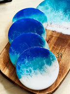 A set of ocean-themed resin coasters in shades of blue and white, resembling waves. The coasters are round and sit on a wooden cutting board. The image also includes a larger, matching serving tray with a similar design.