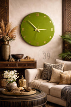 A large, round, olive green wall clock with gold-colored hands and Roman numerals. The clock is mounted on a neutral-colored wall, with decorative elements like a sofa, pillows, and a coffee table.