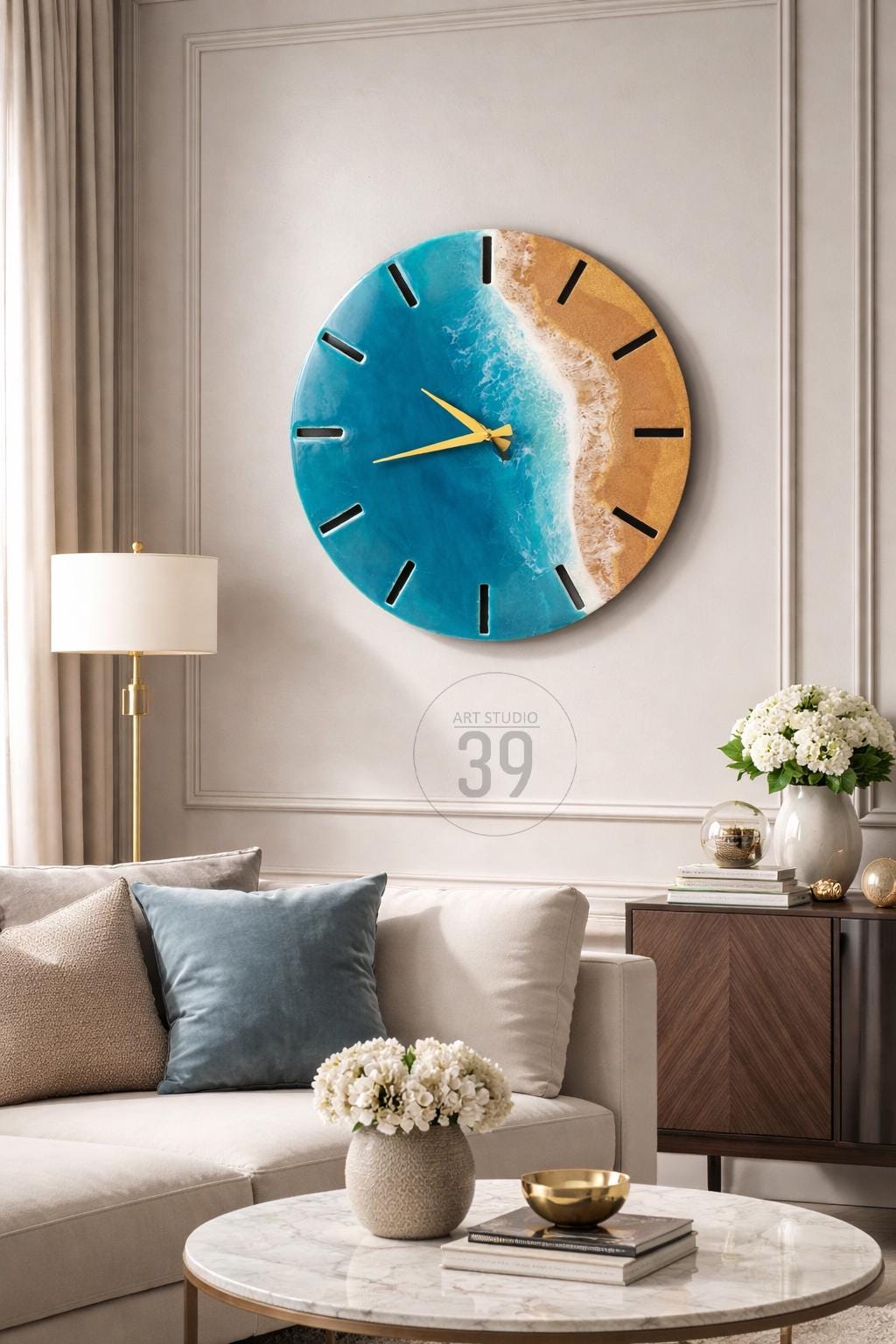 A round wall clock with a beach-themed design. The clock face features a blue ocean, a sandy shore, and gold-colored hands. The clock is mounted on a light-colored wall. The text "ART STUDIO 39" is in the center.