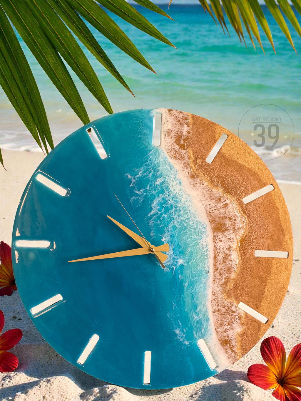 A round wall clock with a beach-themed design. The clock face features a blue ocean and sandy beach design. The clock has gold-colored hands and white rectangular hour markers. The text "ART STUDIO 39" is visible on the clock face.