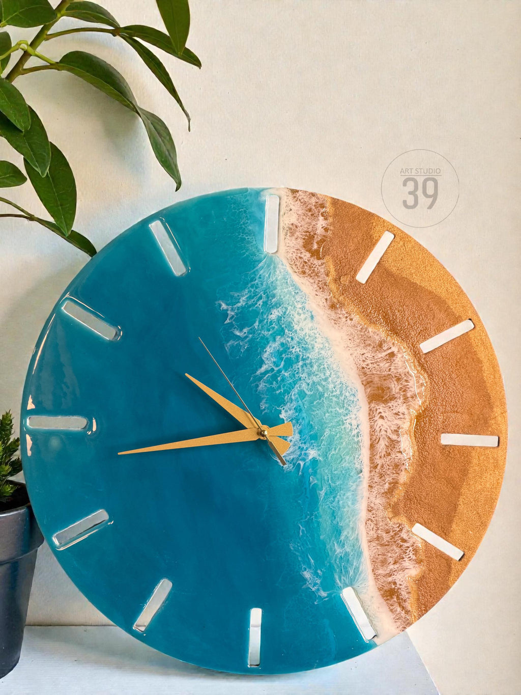 A round wall clock with a beach-themed design. The clock face features a blue ocean, white foamy waves, and a golden-colored beach. The clock has gold-colored hands and white rectangular hour markers. The text "ART STUDIO 39" is visible.