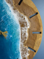 A round wall clock with a beach scene design. The clock face features a golden sand-like texture, blue ocean waves, and gold-colored clock hands. The clock has black rectangular hour markers.