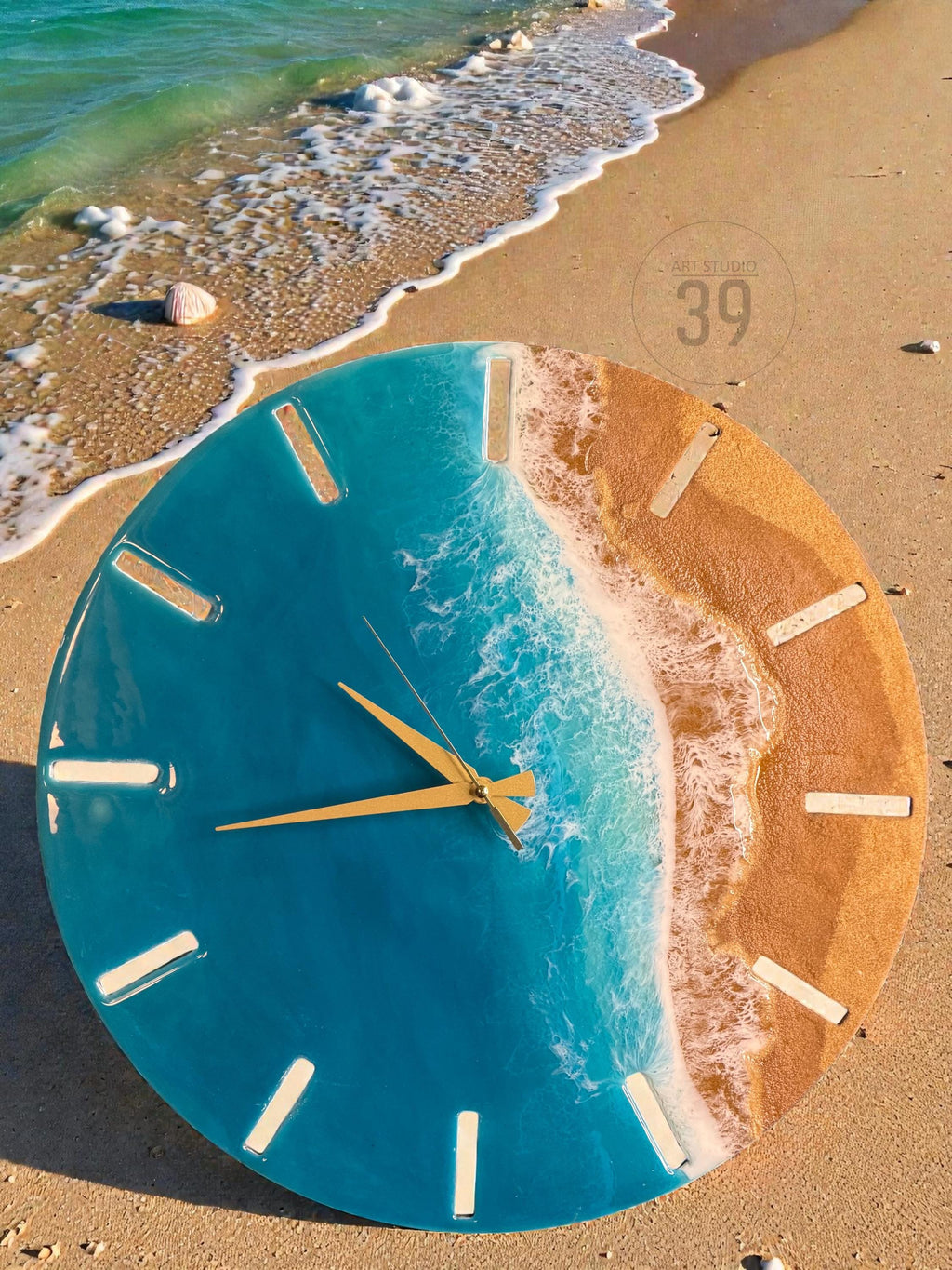 A round wall clock with a beach-themed design. The clock face features a blue resin ocean with white wave details and a sandy beach section. Gold-colored clock hands and white hour markers complete the design. The clock is on a sandy surface.