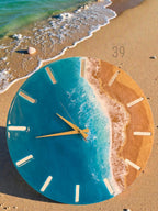 A round wall clock with a beach-themed design. The clock face features a blue resin ocean with white wave details and a sandy beach section. Gold-colored clock hands and white hour markers complete the design. The clock is on a sandy surface.