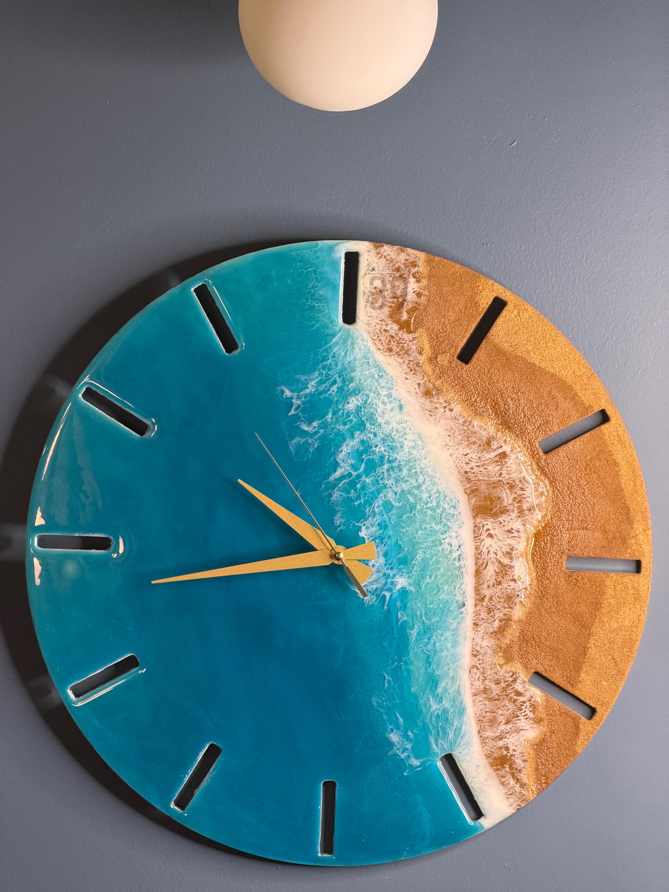 Round wall clock with a beach scene design. The clock face features a blue ocean, a sandy beach, and gold-colored clock hands. The clock has a glossy finish and a modern aesthetic.
