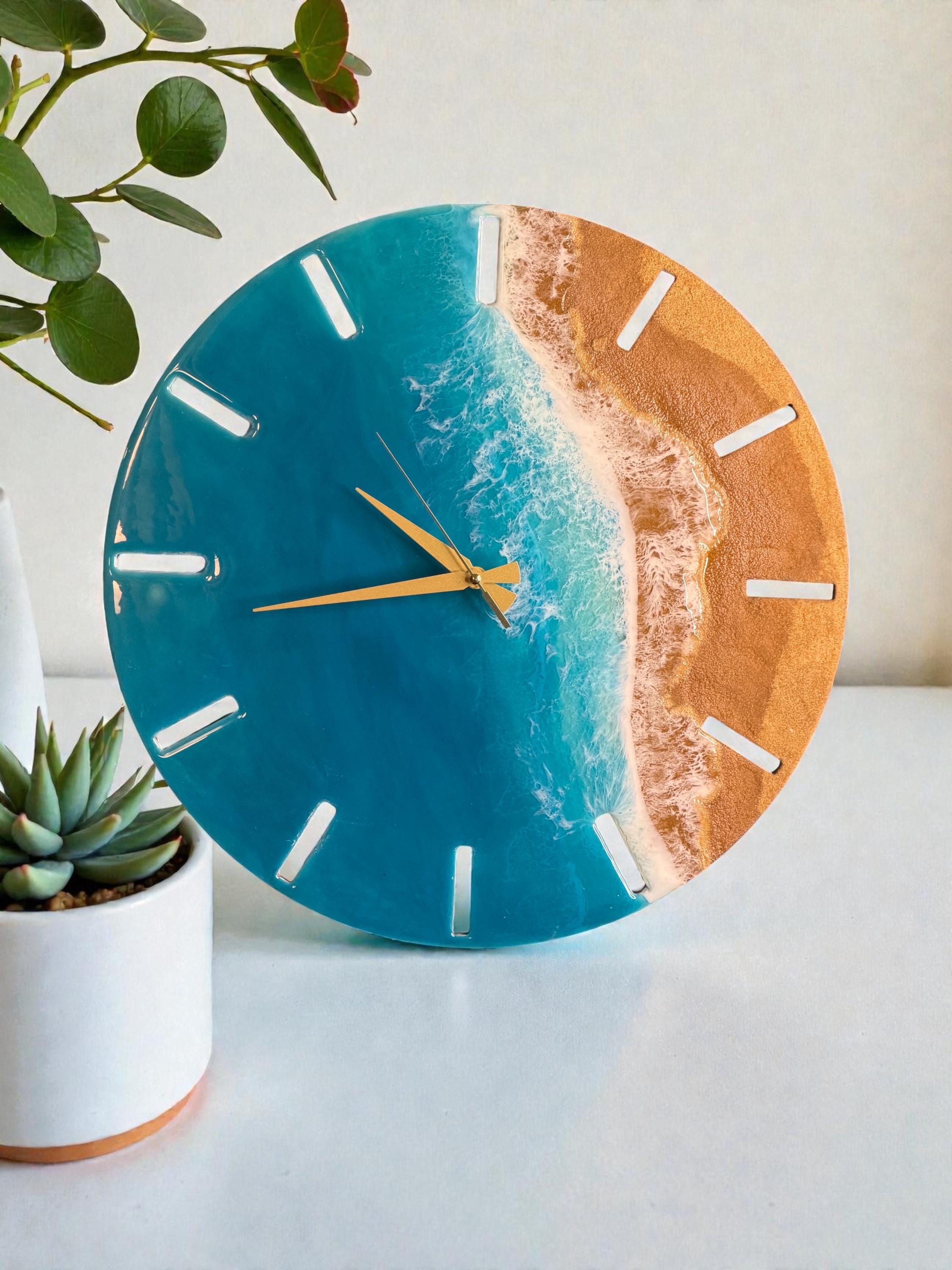 Round wall clock with a beach scene design. The clock face features a blue ocean, white waves, and a golden sand beach. The clock has gold-colored hands and white markers. The clock is approximately 10 inches in diameter.