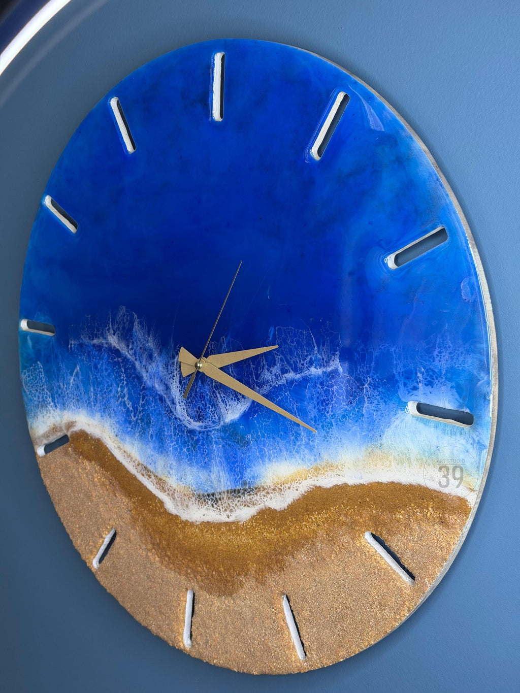 Round wall clock with a beach-themed design. The clock face features a blue ocean and golden sand, with white foamy waves. The clock has gold hands and white markers. The number "39" is visible on the clock face.