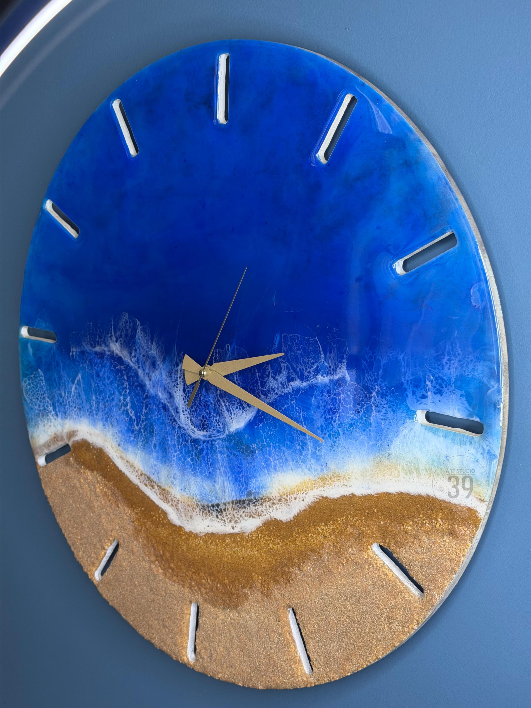 Round wall clock with a beach-themed design. The clock face features a blue ocean and golden sand, with white foamy waves. The clock has gold hands and white markers. The number "39" is visible on the clock face.