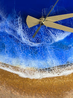 A wall clock with a beach scene design. The clock face features a blue ocean with white foamy waves and a sandy shore. The clock hands are gold-colored.