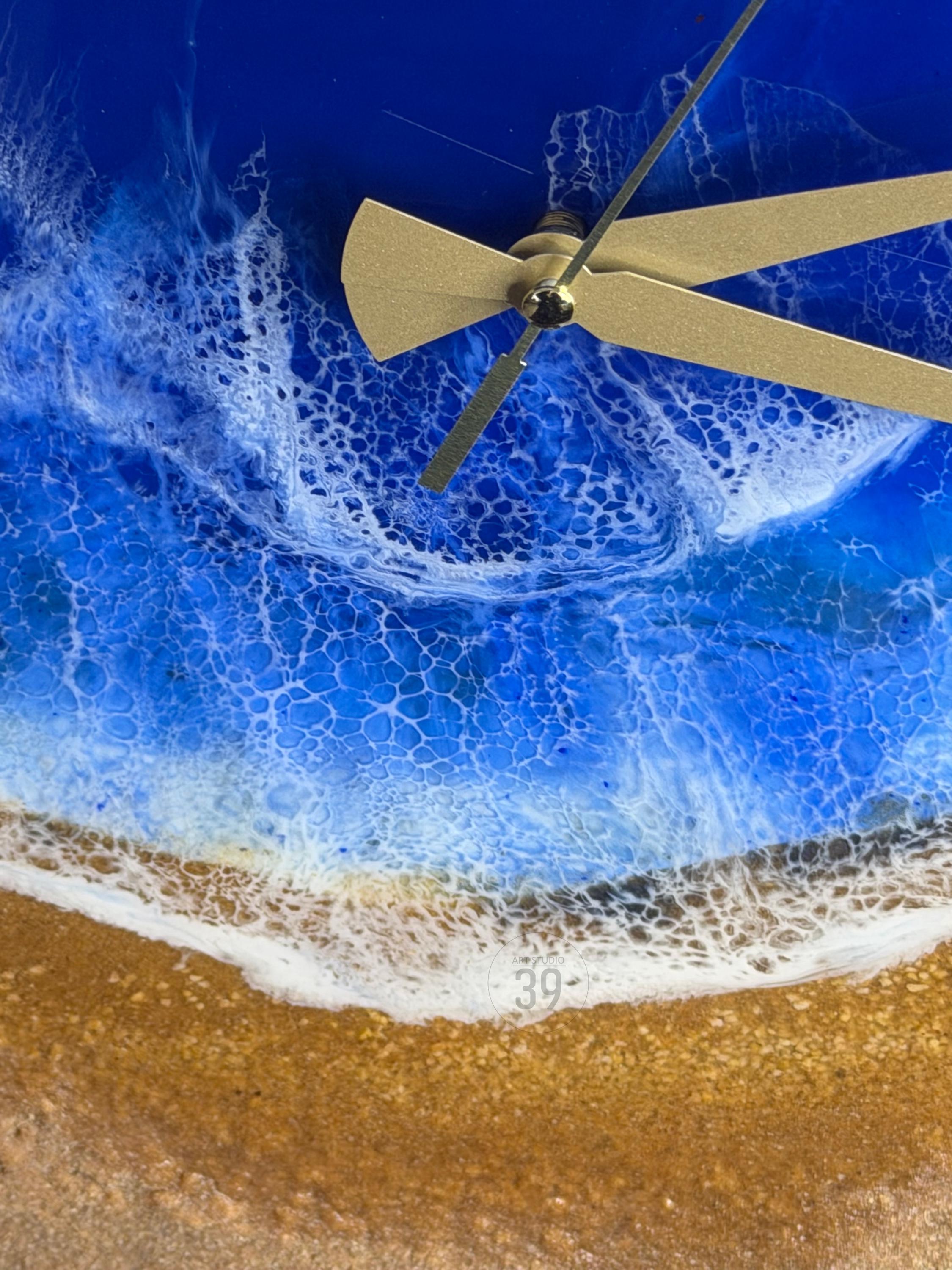 A wall clock with a beach scene design. The clock face features a blue ocean with white foamy waves and a sandy shore. The clock hands are gold-colored.