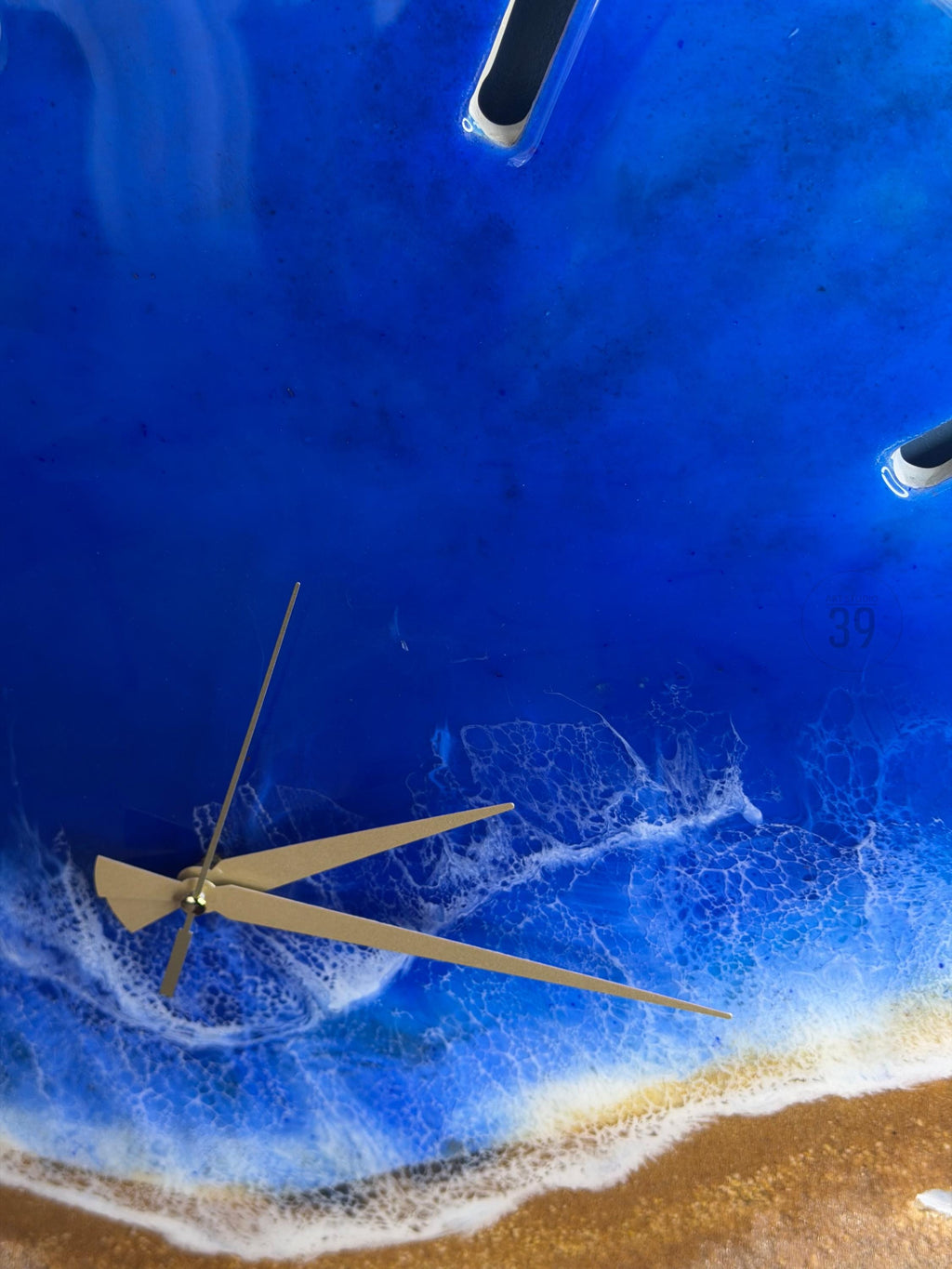 A round wall clock with a beach-themed design. The clock face features a deep blue resin representing the ocean, with white foamy waves and a sandy shore. Gold-colored clock hands and a partial view of the clock's frame are visible.