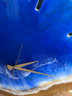 A round wall clock with a beach-themed design. The clock face features a deep blue resin representing the ocean, with white foamy waves and a sandy shore. Gold-colored clock hands and a partial view of the clock's frame are visible.