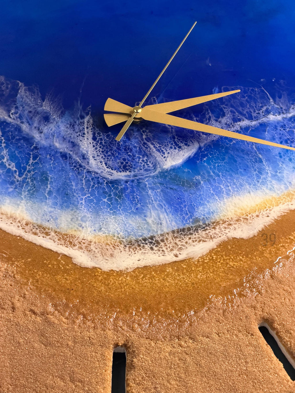 A round wall clock with a beach scene design. The clock face features a blue ocean with white waves and a sandy beach. Gold-colored clock hands indicate the time. The clock has a glossy finish.
