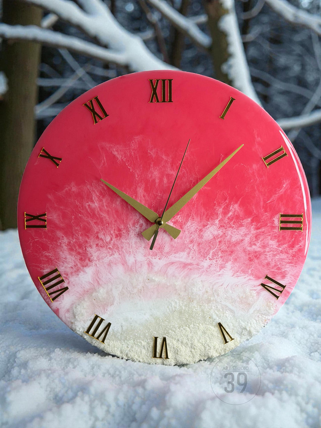 A round wall clock with a pink and white resin design. The clock face features gold Roman numerals and hands. The clock is sitting in the snow. The text "ART STUDIO 39" is visible.