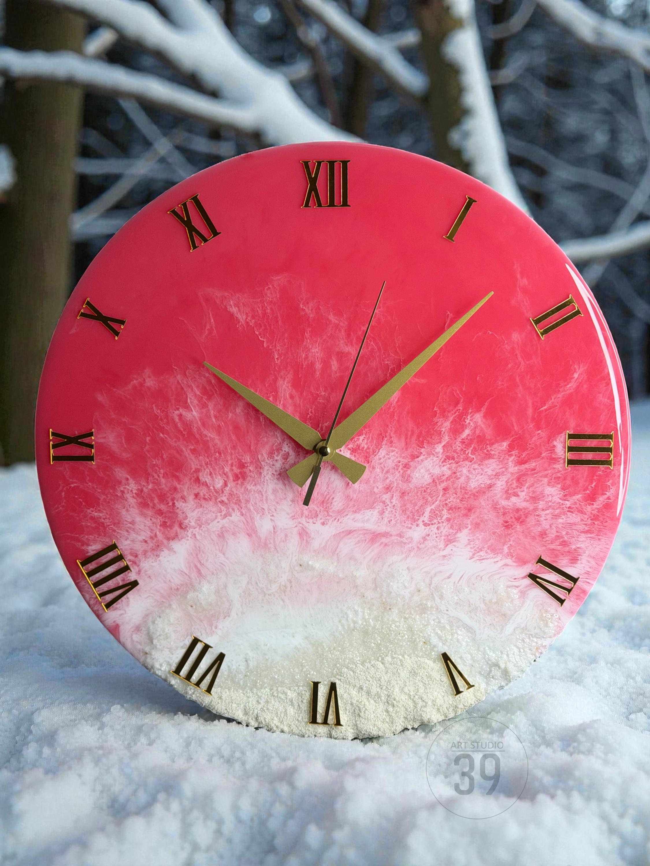 A round wall clock with a pink and white resin design. The clock face features gold Roman numerals and hands. The clock is sitting in the snow. The text "ART STUDIO 39" is visible.