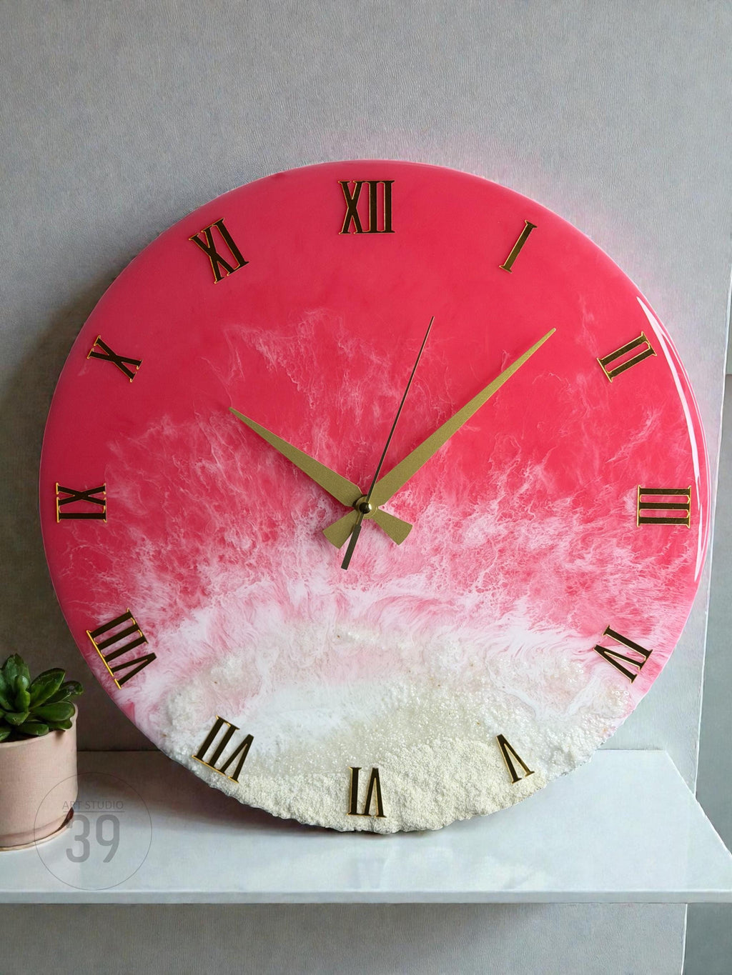 A round wall clock with a vibrant pink and white resin design. The clock features gold Roman numerals and hands. The bottom portion has a textured, sandy appearance. The clock is a decorative piece for home decor.