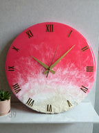 A round wall clock with a vibrant pink and white resin design. The clock features gold Roman numerals and hands. The bottom portion has a textured, sandy appearance. The clock is a decorative piece for home decor.