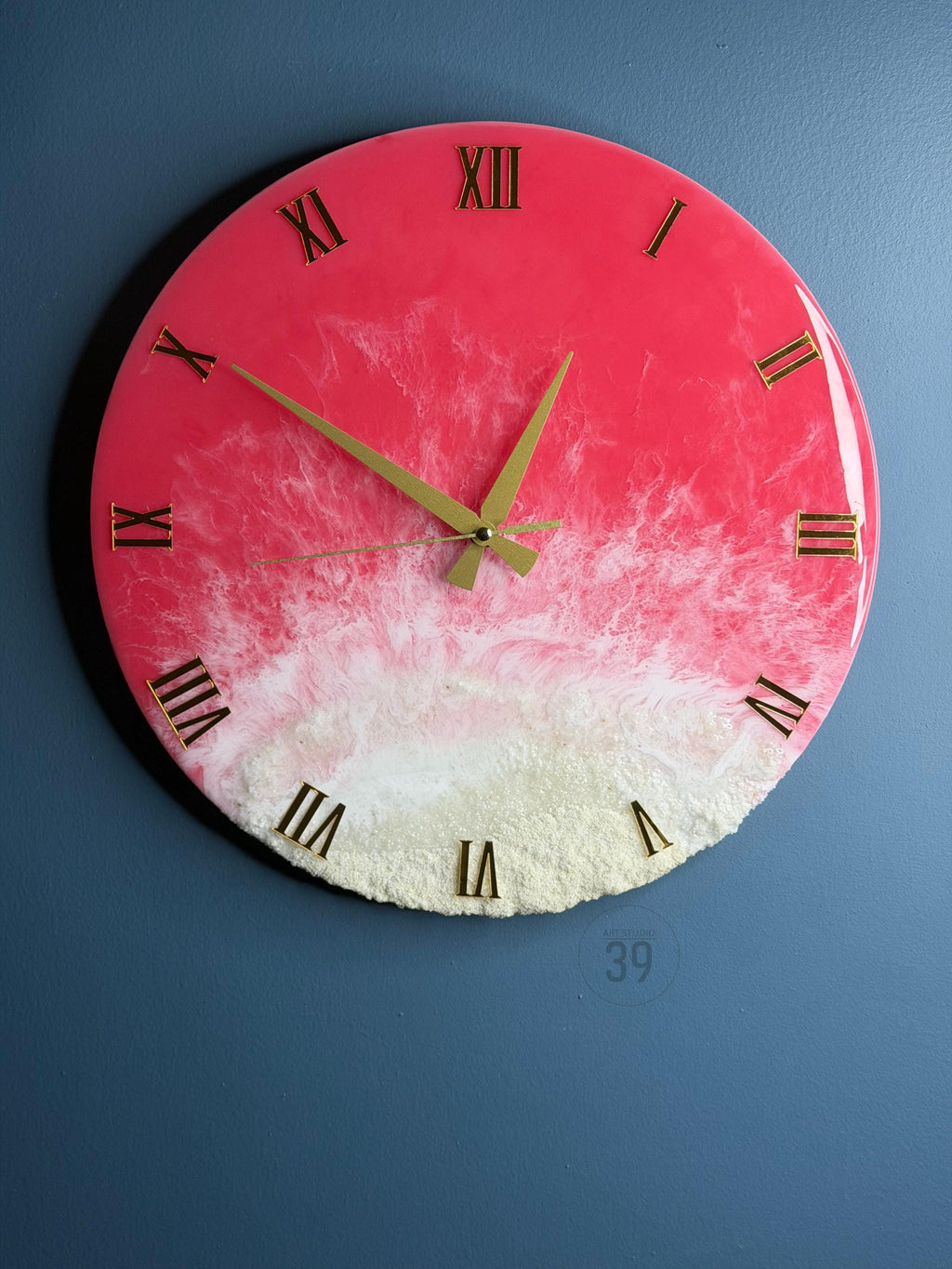 A round wall clock with a pink and white resin design. The clock features gold Roman numerals and hands. The bottom portion has a textured, sandy appearance. The clock is mounted on a blue wall.