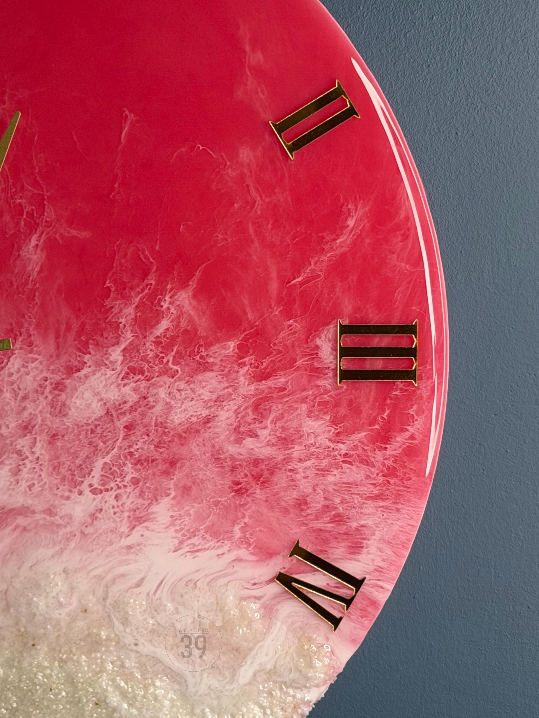 A close-up of a round wall clock with a vibrant pink and white resin design. The clock features gold Roman numerals and hands. The number "39" is visible near the bottom. The clock is a decorative home decor item.