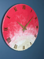 A round wall clock with a pink and white resin design. The clock features gold Roman numerals and hands. The bottom portion has a textured, sandy appearance. The number "39" is visible near the bottom.