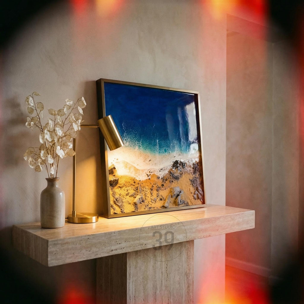 Square framed artwork depicting a beach scene with blue ocean, white waves, and golden sand. The artwork is displayed on a stone shelf, next to a gold desk lamp and a vase with decorative stems. The artwork is framed in a gold frame.