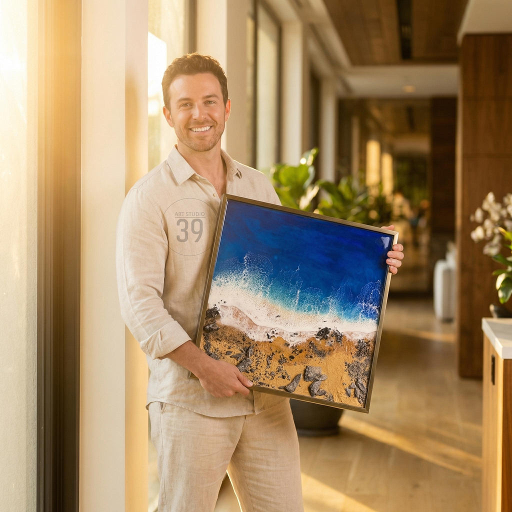 A framed artwork depicting a coastal scene with a vibrant blue ocean, white foamy waves, and a sandy beach with rocks. The artwork is held by a person wearing a beige shirt and pants.