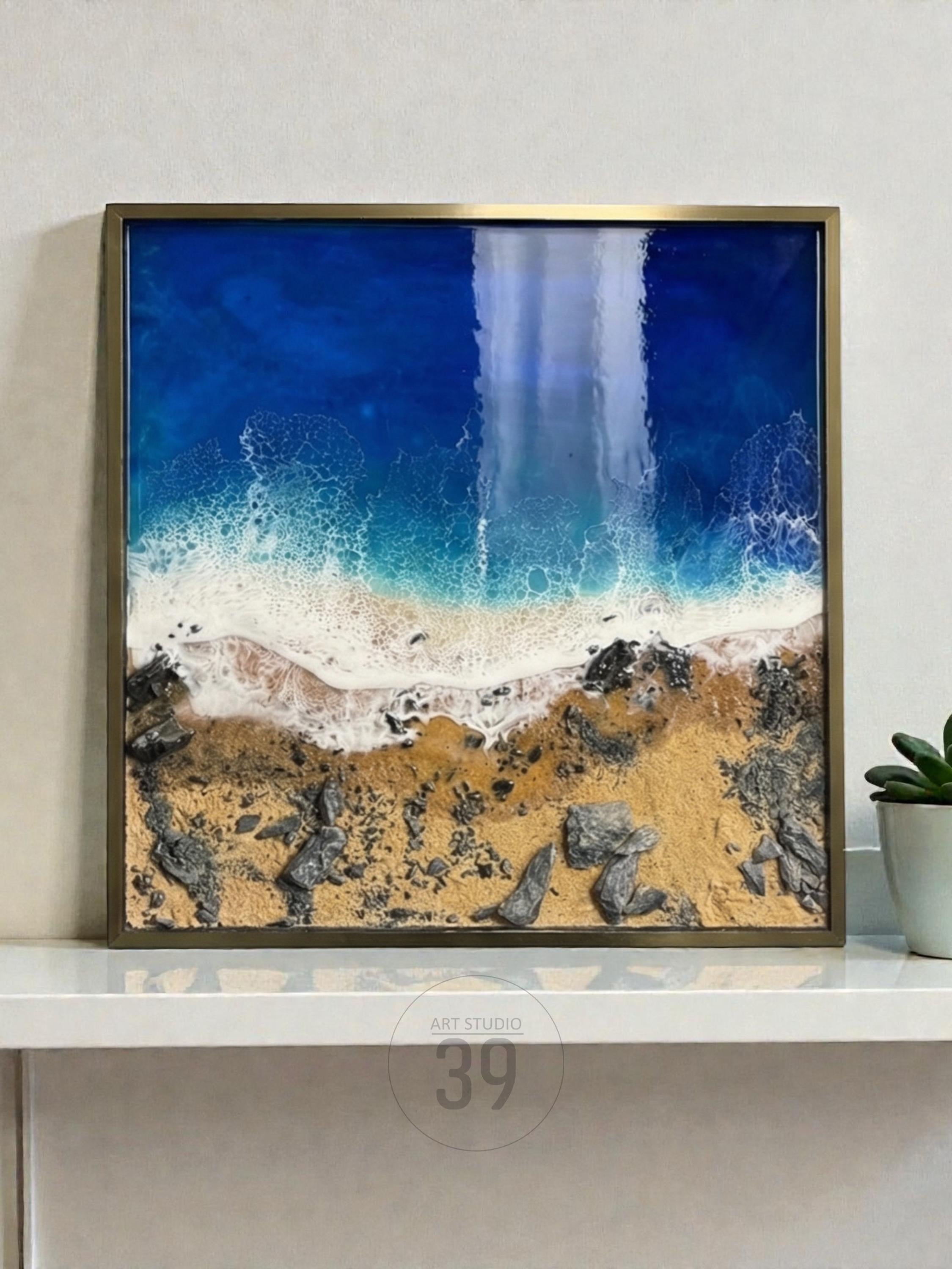 Square framed artwork depicting a coastal scene. The top portion shows deep blue ocean, transitioning to white foamy waves, a sandy beach, and rocky terrain. The artwork is framed in a gold-colored metal frame. The text "ART STUDIO 39" is visible.
