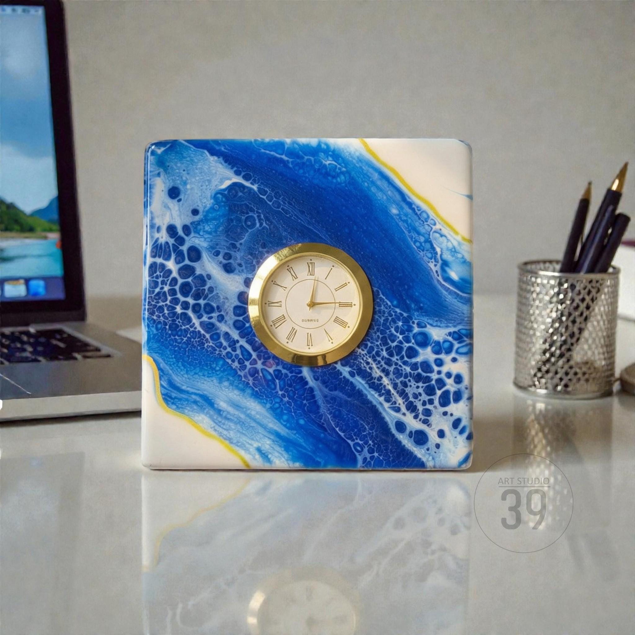 Square desk clock with a gold-tone clock face and Roman numerals. The clock's face is set in a square block with a blue and white abstract design. The clock is on a white surface with a laptop and pen holder in the background.