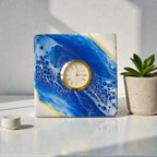 Square clock with a gold-tone bezel and a cream-colored face. The clock is set into a square block with a blue and white abstract design. A small potted plant and a white ceramic piece are next to the clock.
