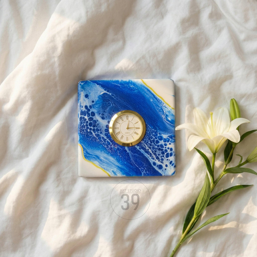 Square decorative clock with a gold-tone bezel and a white face, set on a blue and white abstract art background. A white lily flower and the text "ART STUDIO 39" are also in the image.