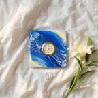 Square decorative clock with a gold-tone bezel and a white face, set on a blue and white abstract art background. A white lily flower and the text "ART STUDIO 39" are also in the image.